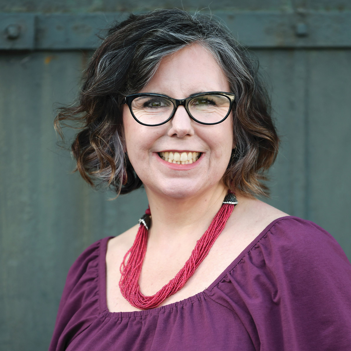 Lorraine Besser color portrait. Lorraine is a older, white woman with short chin-length wavy hair in silver and brown. She posed outside framed by dark green barn doors. She is wearing a red beaded necklace and a purple top.