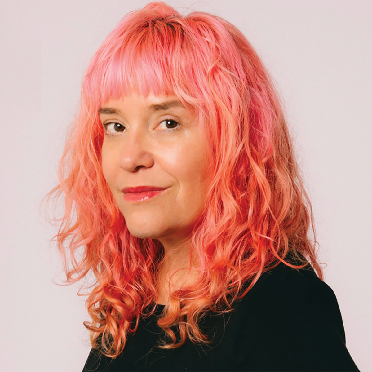 Michelle Tea color portrait. Michelle is smiling and posed against a neutral background. Michelle has soft pink wavy hair and is wearing a dark-colored top.