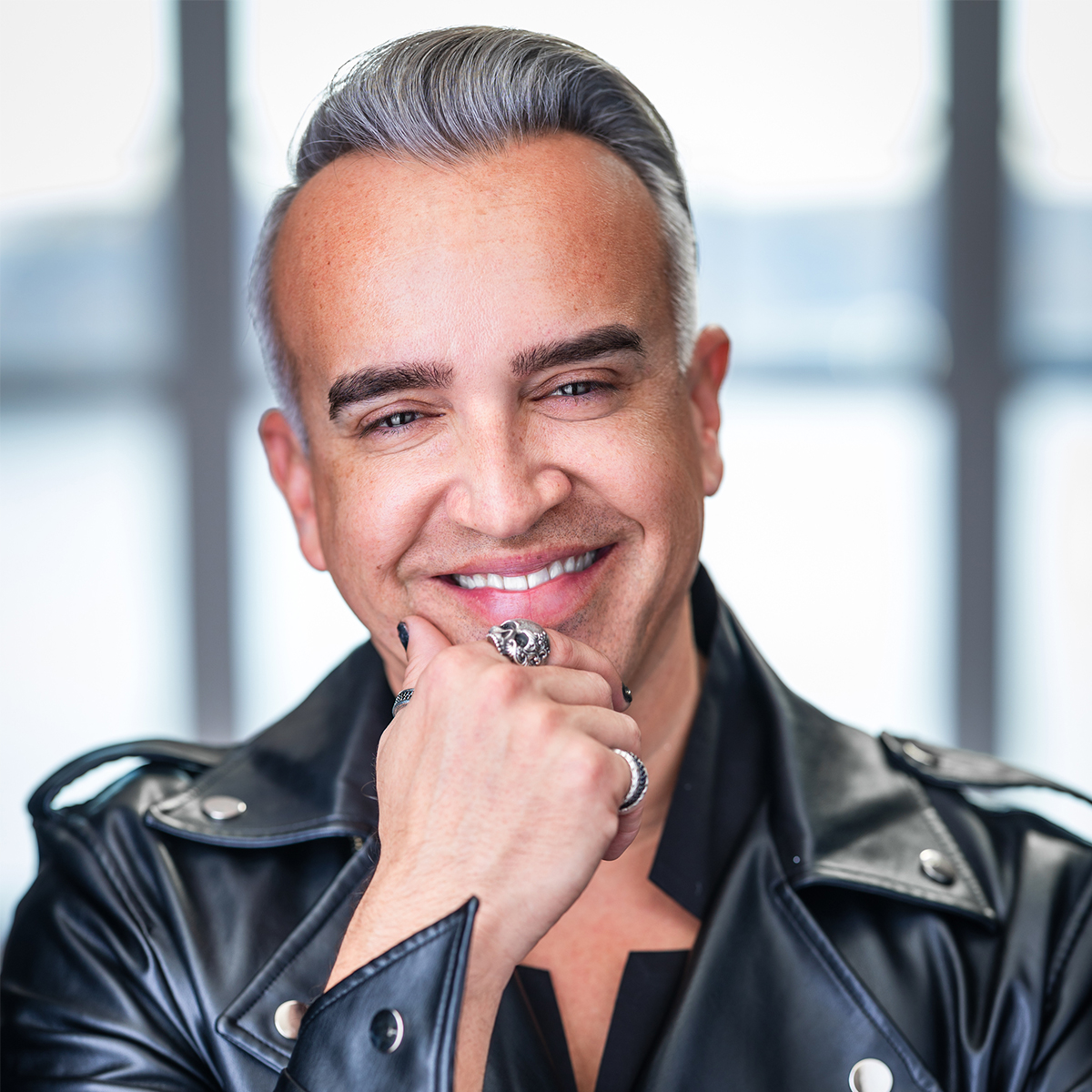 Dr. Roger Kuhn color portrait. Dr. Kuhn is a Two-Spirit Indigequeer person who is posed in front of blurred windows, is smiling, and and has one hand resting on his chin. He is wearing a black leather jacket, a silver ring, his nails are painted a dark-color.