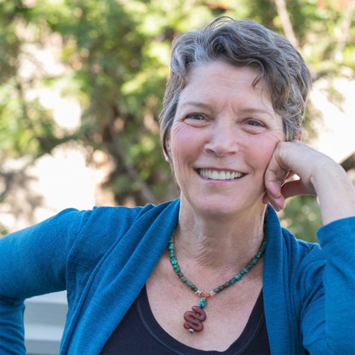 Peggy Martin color portrait. Peggy is an older, white woman, who is posed outside, smiling, and has her cheek resting on one hand. She is wearing a turquoise cardigan, a black top, and a beaded necklace.
