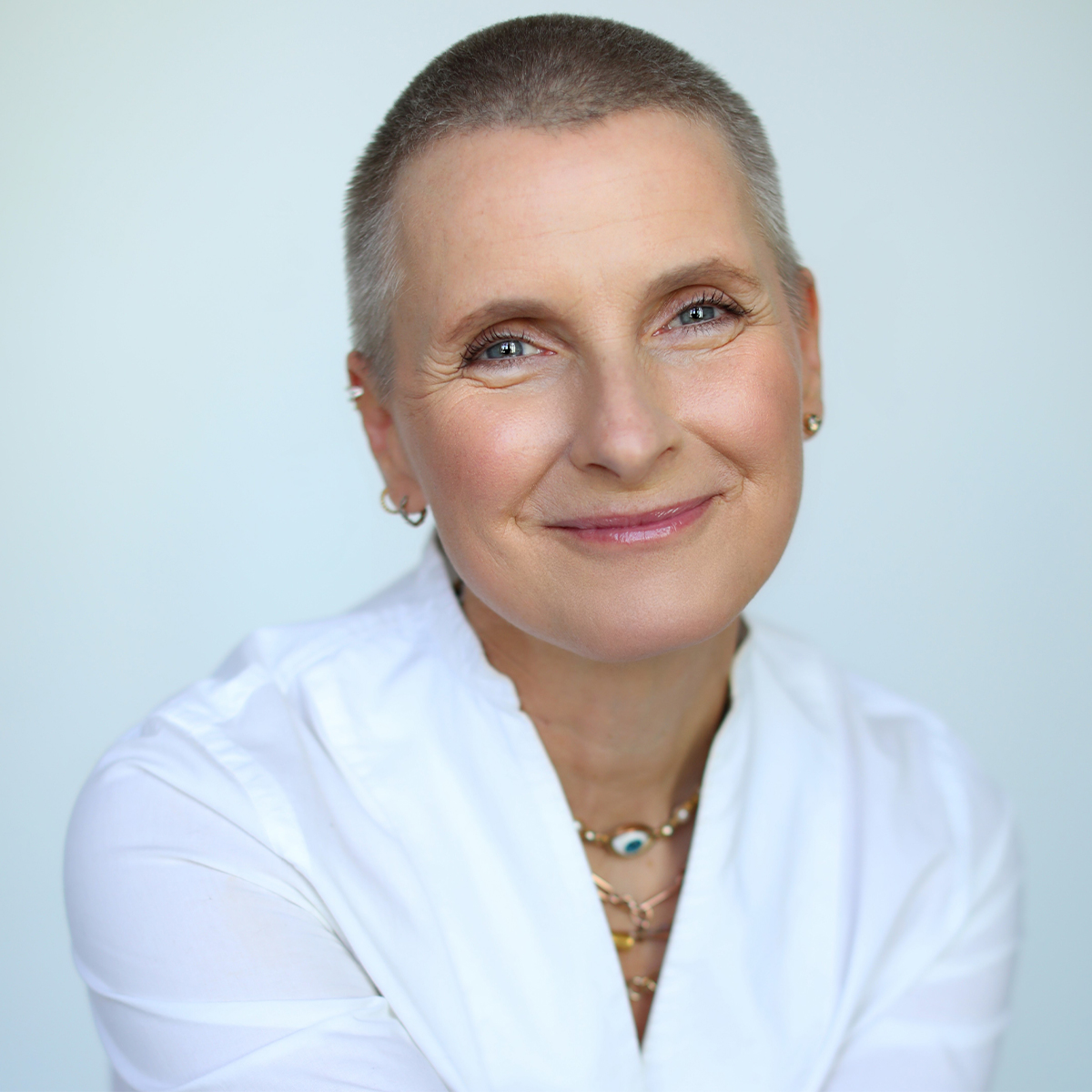 Elizabeth Gilbert color portrait. Elizabeth is smiling, has a shaved head, and is wearing a white top with layers of necklaces.