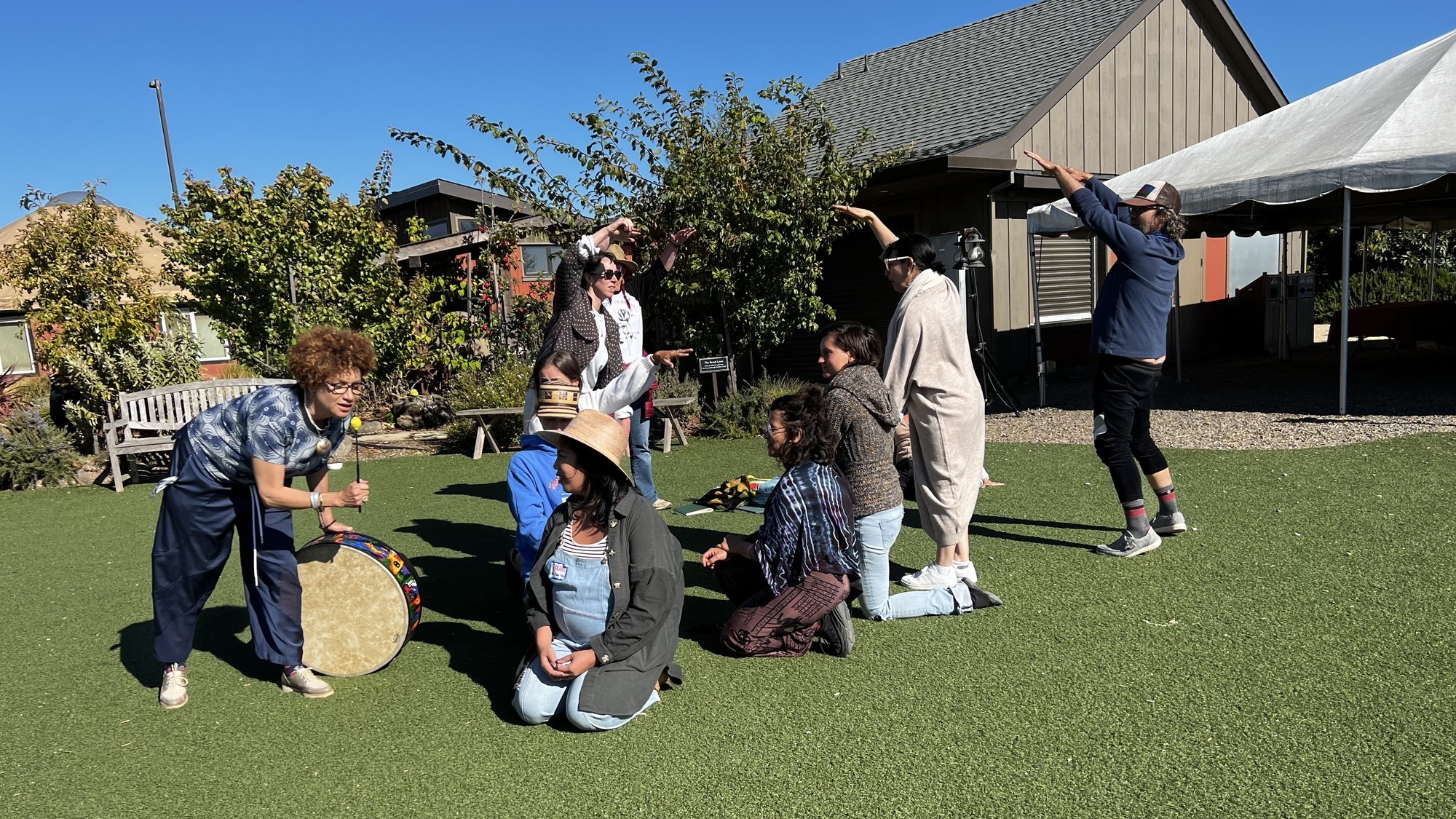 Photo of a group of students in animated motion performing outdoors with a drum. One woman is holding a drumstick like a mic.