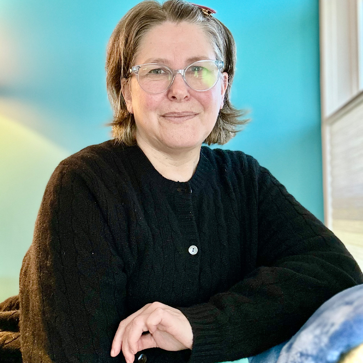 Rebecca Bloom color portrait. Rebecca is wearing thin, clear glasses, has short, light-colored hair pinned back from her smiling face. She is waring a dark-colored cardigan and has one arm resting on the arm of a chair.