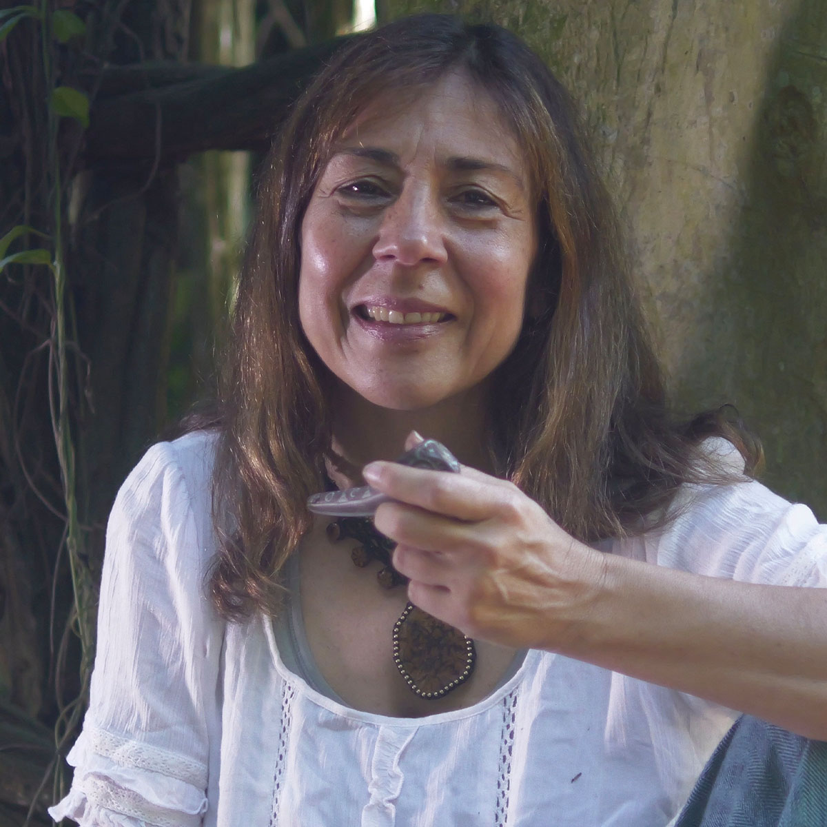 Susana Bustos color portrait. She is seated outside, holding a carved item in her hand, and is wearing a cotton blouse, a large wooden necklace, and is smiling.