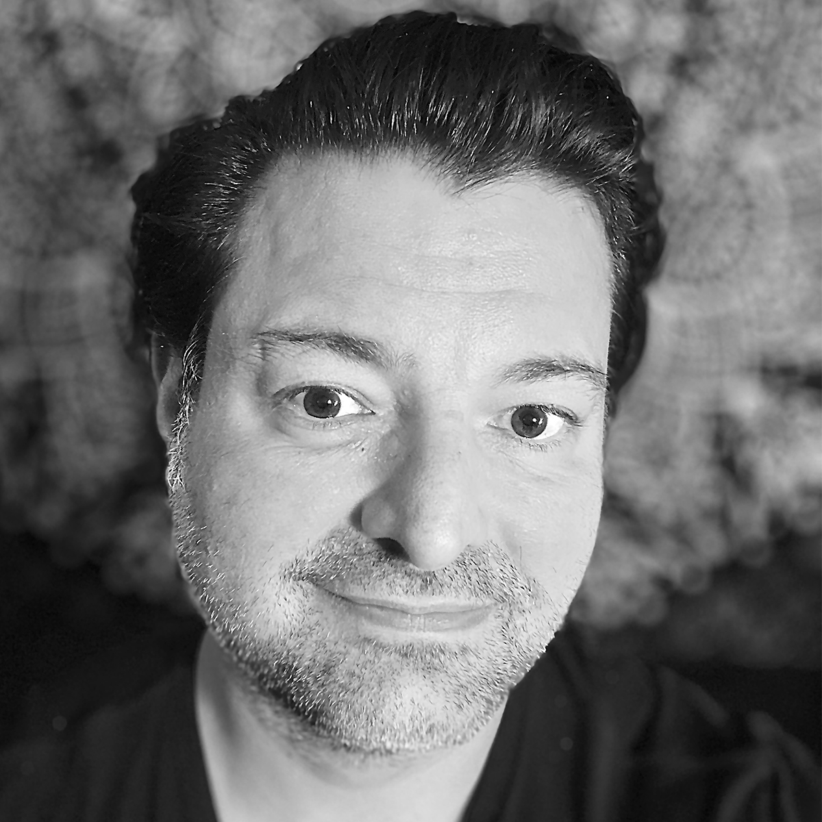 Ralph De La Rosa black and white portrait. Ralph is smiling in this portrait, has short, slick-backed hair.