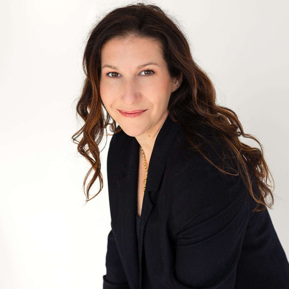 Britt Frank color portrait. Britt is a white person with long brown, wavy hair that ends past her shoulders. She is smiling and is wearing a black suit.