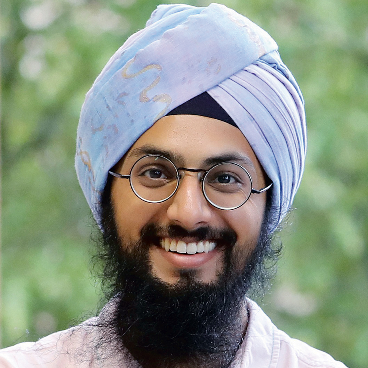 Manvir Singh color portrait. Manvir is wearing a light blue and gray silken head wrap, round glasses, and has a beard that ends past his chin. He is posed outside and is smiling.