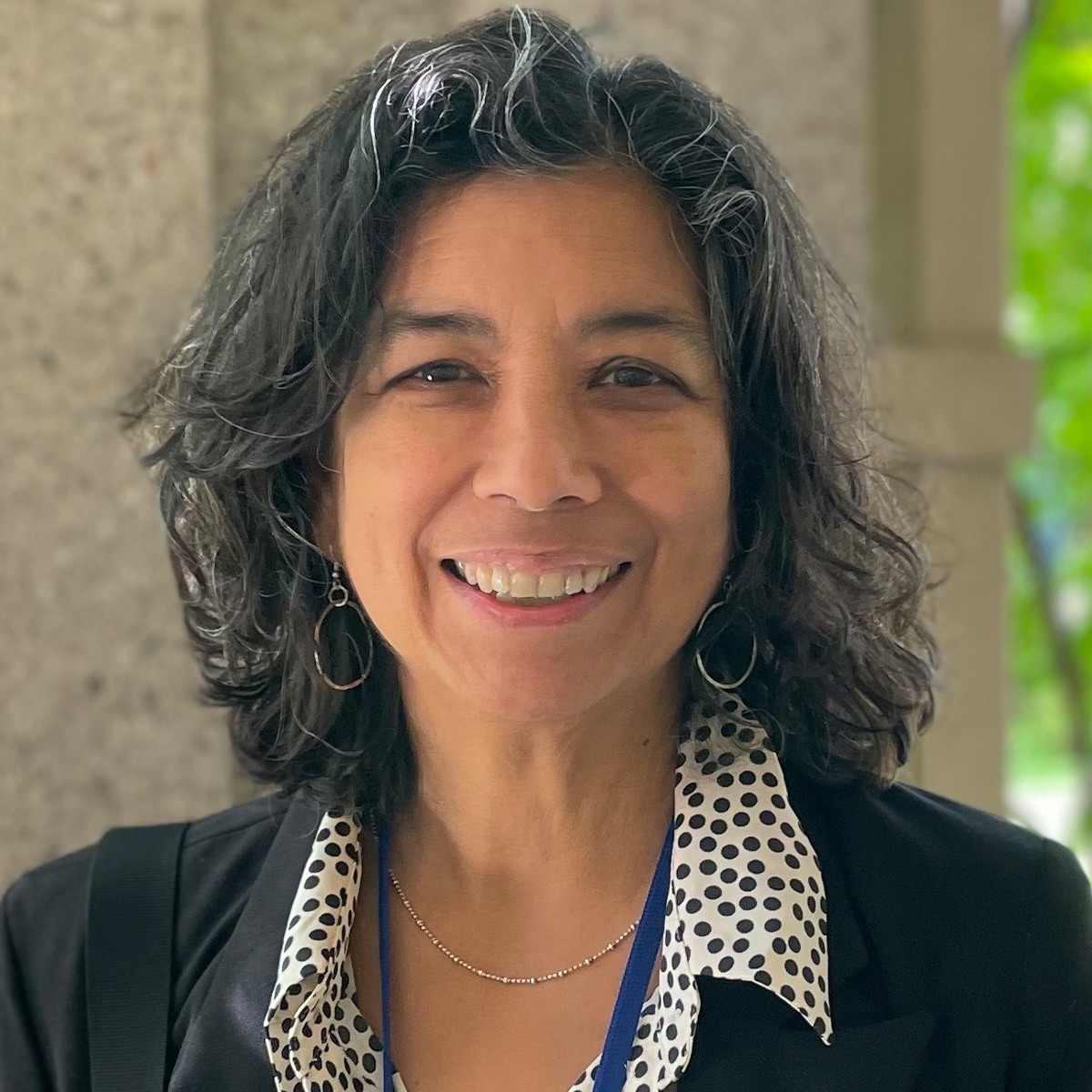Licia Sky color portrait. Licia is smiling in this portrait has dark-colored medium-length wavy hair, has light-brown skin, and is wearing a dark-colored jacket and polka-dotted top.