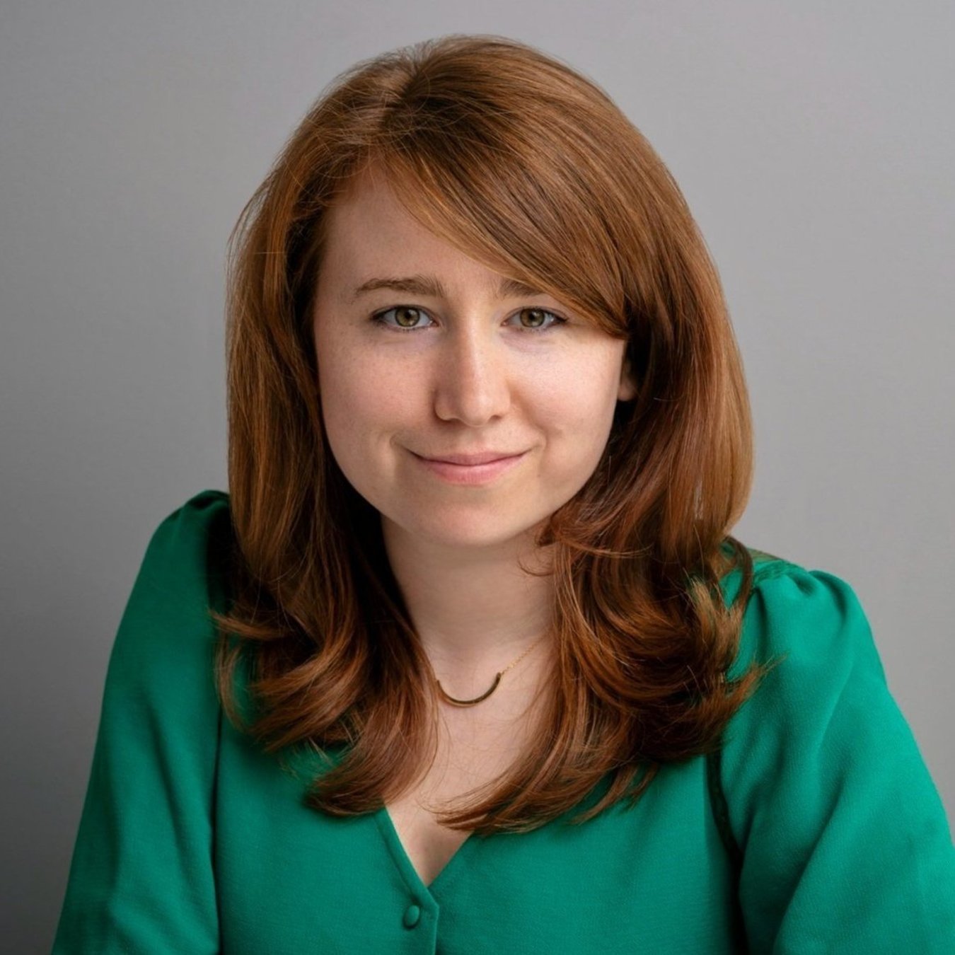 Rhaina Cohen color portrait. Rhaina is a white person with slightly wavy brown hair and is wearing a emerald green top and smiling.