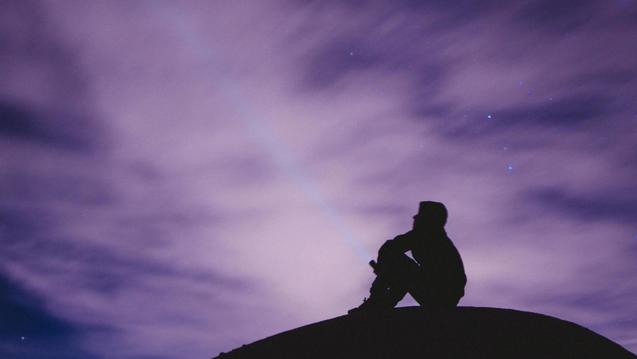 Photo of a silhouetted person sitting on a hill with a starry purple sky behind.