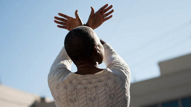 Photo of a Black person facing away from camera with hands raised to form a bird silhouette against a blue sky