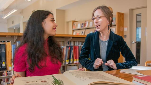 Photo of two women talking in a library with a table full of open books