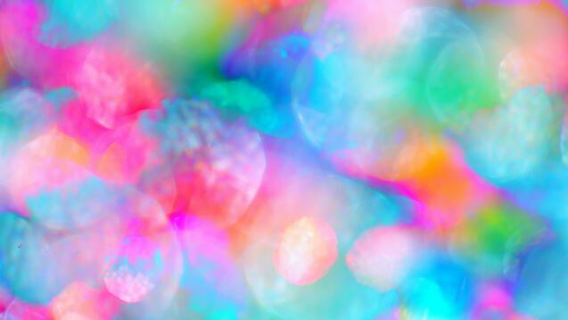 A vibrant and abstract blend of multicolored blurred lights in shades of pink, blue, green, and orange, creating a soft, dreamlike bokeh effect.