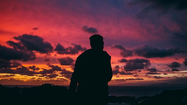 A silhouette of a person standing against a striking sunset with colorful clouds. The sky is filled with shades of deep pink, purple, and orange, creating a dramatic scene. The person appears to be gazing at the horizon, possibly overlooking a body of water, with clouds scattered across the sky. The overall mood of the image is serene and contemplative.