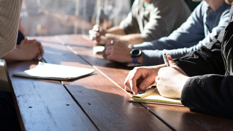 Close up photo of a group of hands writing on pads of paper at a wood table