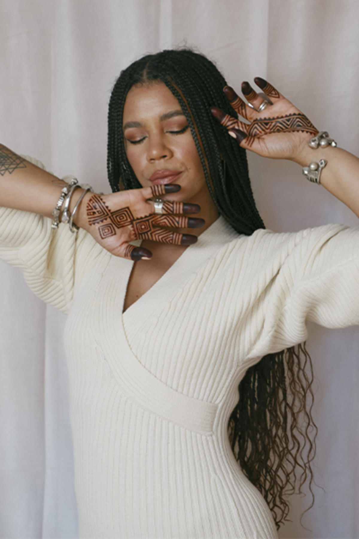 Maryam Hasnaa color portrait. Maryam is a Black woman with long, braided hair to her lower back. Her eyes are closed and her arms are raised and hands open across her chin and head.