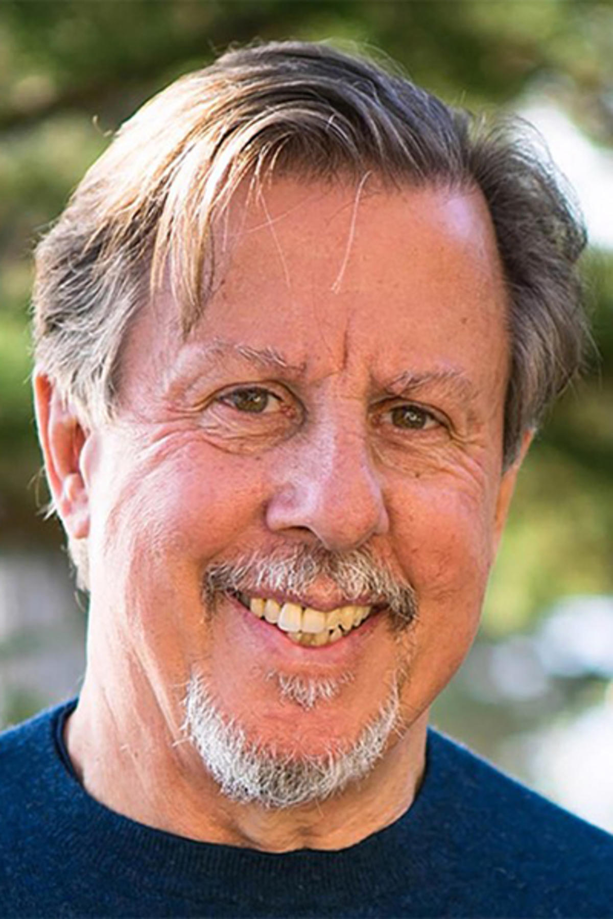 Richard Schwartz color portrait. Richard is an older, white man with silver, brown hair that is short. He is smiling and is posed outside with a tree blurred in the background.
