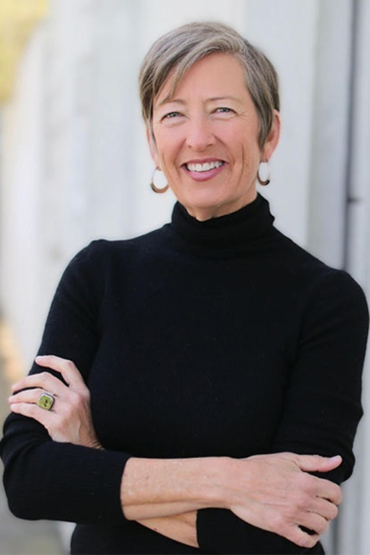 Staci Haines color portrait. Staci is a white woman with short brown, gray hair. Staci is smiling and wears gold hoop earrings and a black turtleneck and her arms are crossed.