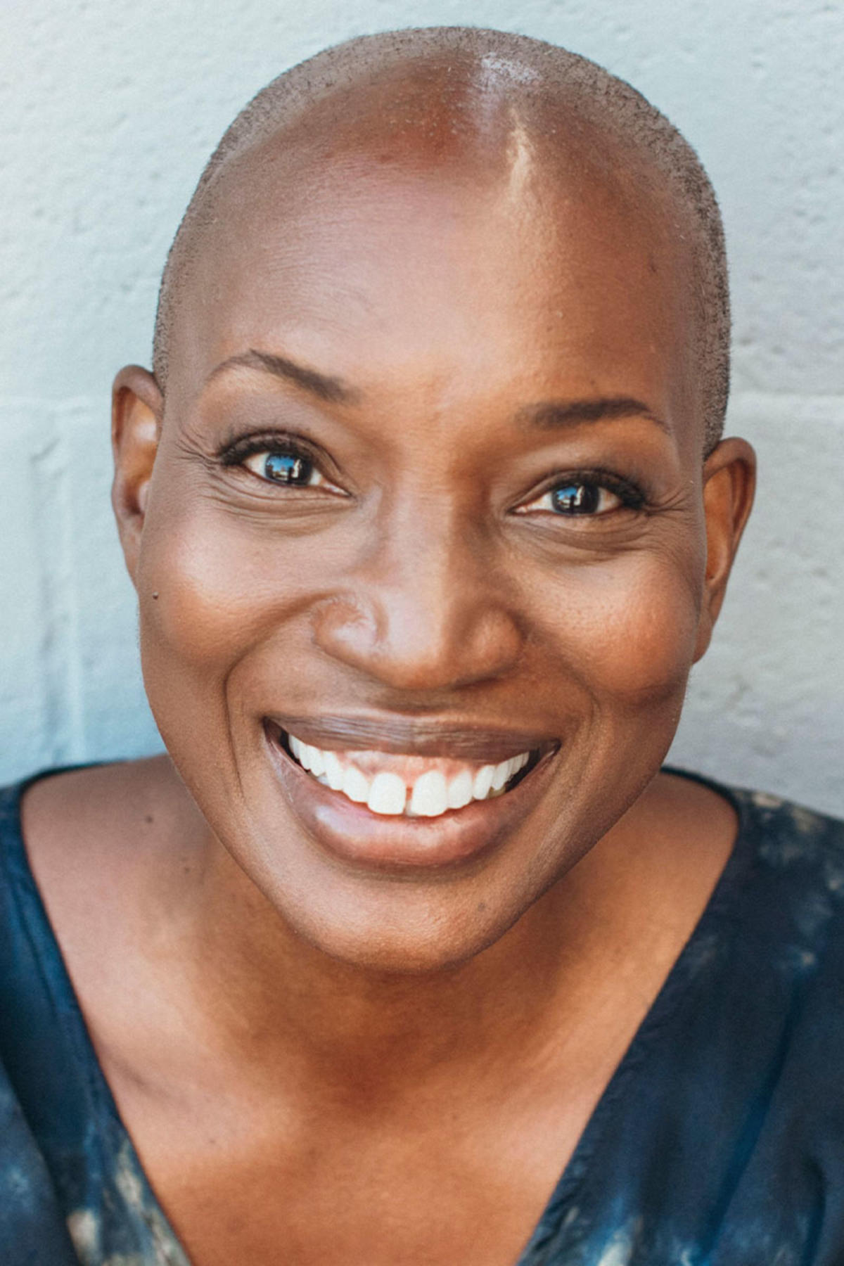 Tracee Stanley color portrait. Tracee is a Black woman leaning against a light-colored wall. Tracee is smiling and wearing a subtly printed top.