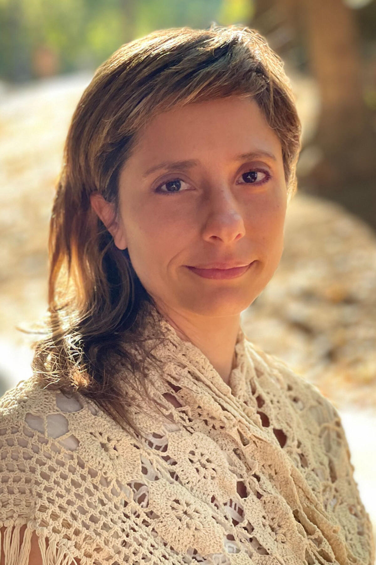 Sara Granovetter color portrait. Sara is a white woman with her hair swept to one side. She is smiling, posed outside, and is wearing a cream knit, lace-line shawl on her shoulders.
