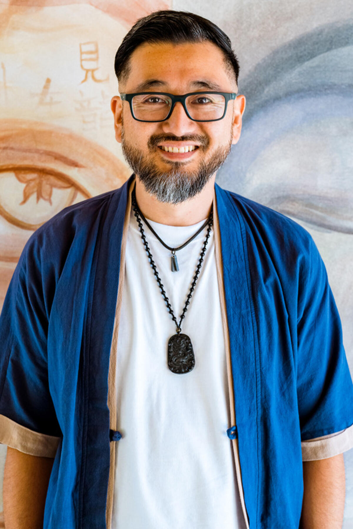 Kazu Haga color portrait. Kazu is a Asian American person who is smiling, while posed standing in front of a mural that is shades of light brown with an open eye painted on the left and shades of blue with a eye closed on the right side.