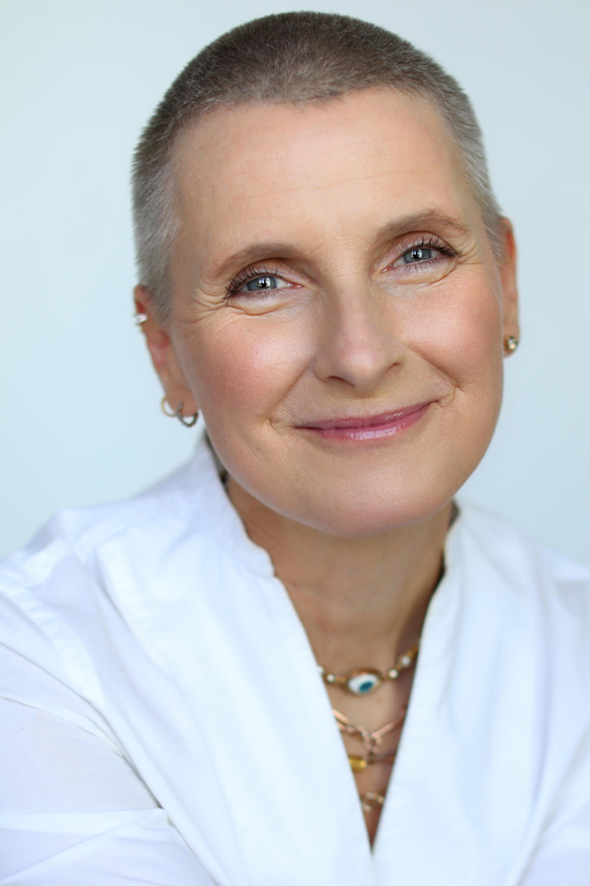 Elizabeth Gilbert color portrait. Elizabeth is smiling, has a shaved head, and is wearing a white top with layers of necklaces.