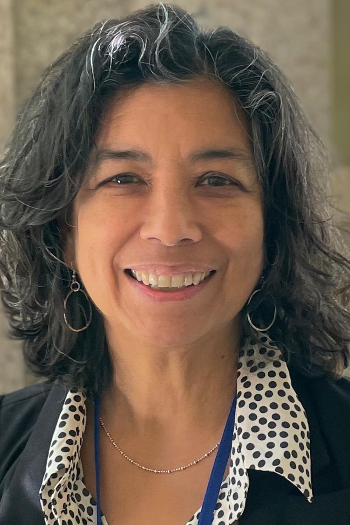 Licia Sky color portrait. Licia is smiling in this portrait has dark-colored medium-length wavy hair, has light-brown skin, and is wearing a dark-colored jacket and polka-dotted top.