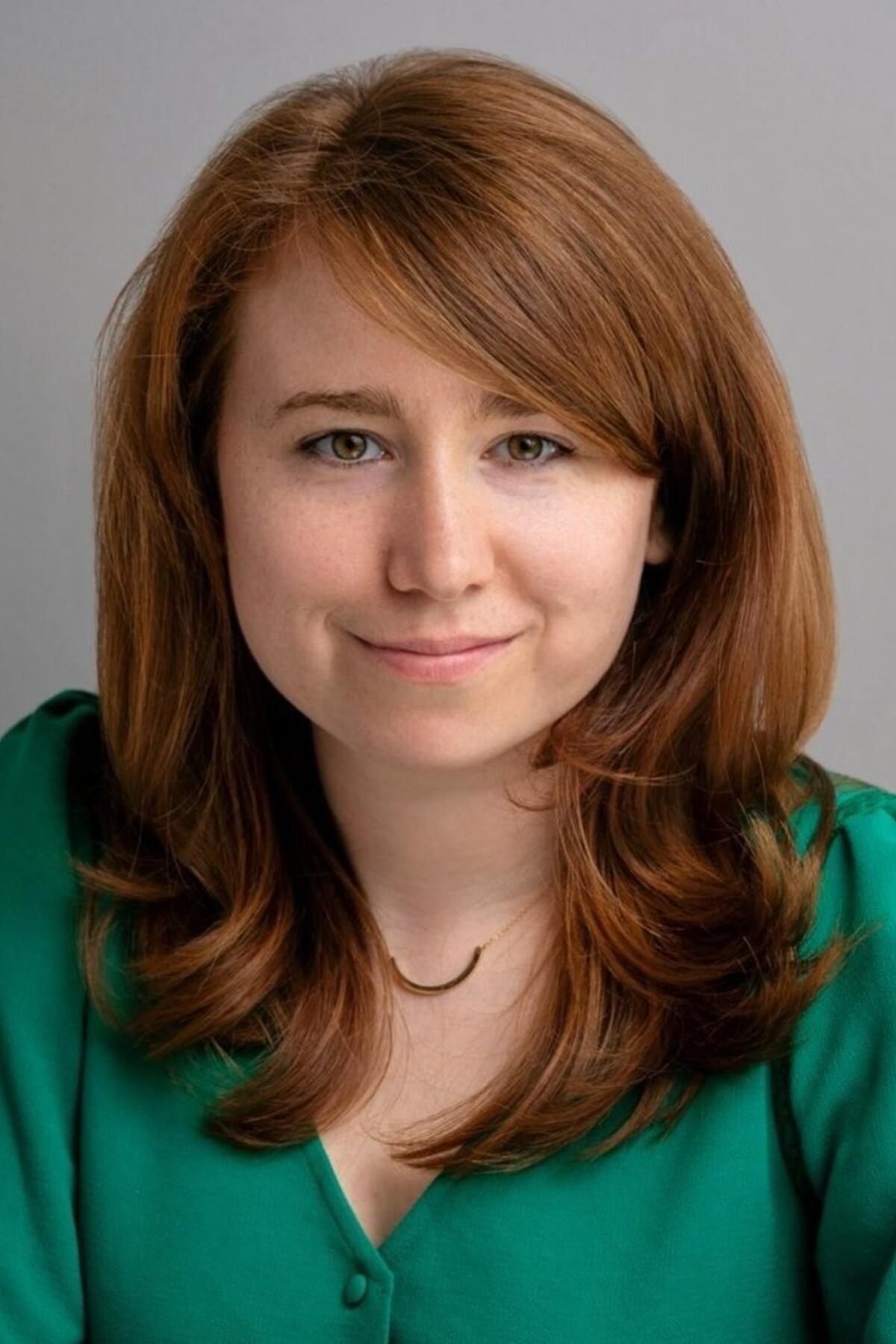 Rhaina Cohen color portrait. Rhaina is a white person with slightly wavy brown hair and is wearing a emerald green top and smiling.