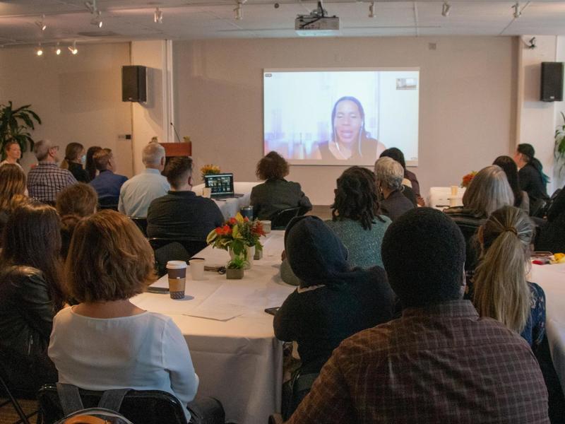 Photo of a large group of seated people looking at an image of a woman projected on a screen